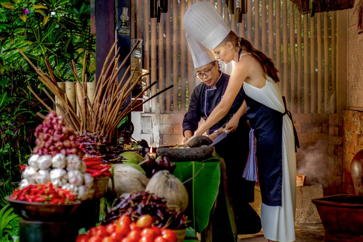Guest joining a Balinese cooking class with a chef, preparing traditional dishes with fresh local ingredients in a tropical open-air kitchen in Ubud, Bali.