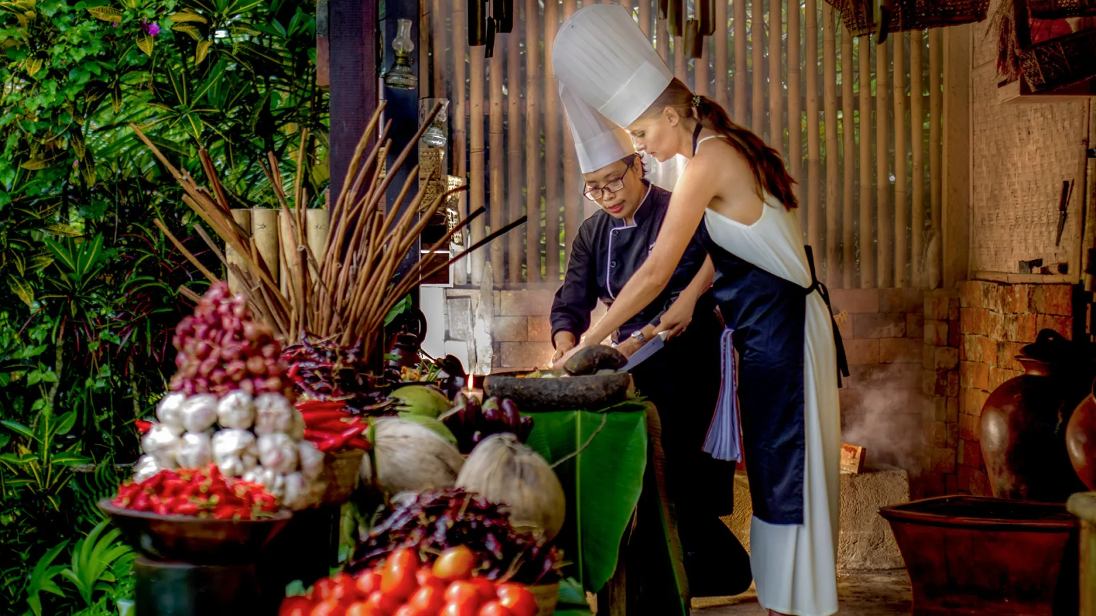 Guest joining a Balinese cooking class with a chef, preparing traditional dishes with fresh local ingredients in a tropical open-air kitchen in Ubud, Bali.
