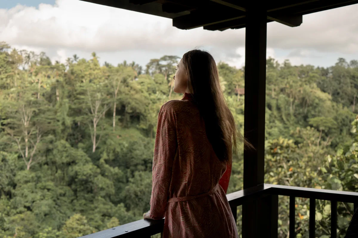 A woman in a pink robe looking out at the lush tropical jungle from a wooden balcony at Nandini Jungle by Hanging Gardens in Bali.